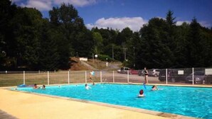 Une piscine avec une terrasse en bois, entourée d’une clôture et bordée d’arbres au loin.