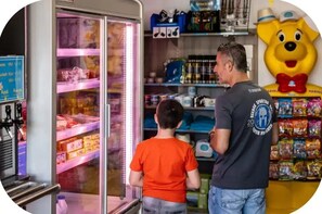 A man and a child in a store with shelves stocked with various products, a large mascot figure, and a refrigerated display case.
