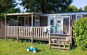 Une maison mobile avec une terrasse, un petit coin cuisine et une voiture jouet sur la pelouse.