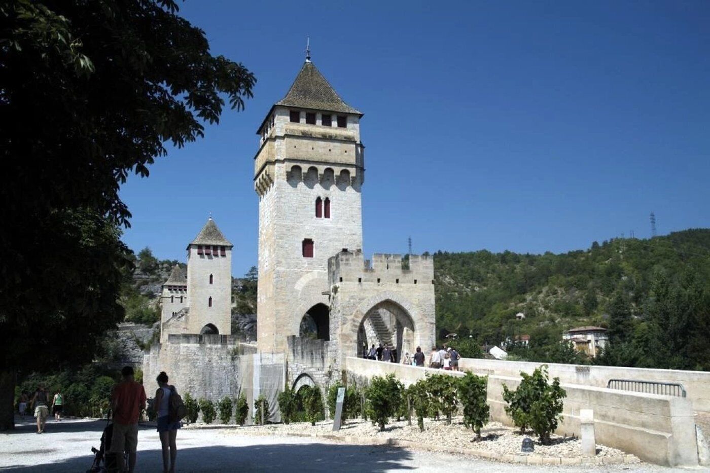 Um castelo de pedra histórico com uma torre central e duas torres menores, rodeado por um espaço pavimentado com árvores e uma ponte.