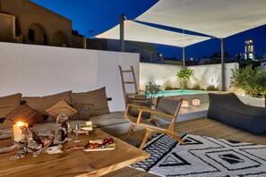 An outdoor seating area with a wooden table, cushions, and a pool. - Arithera Villa in Santorini (Santorini)