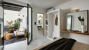 A modern bedroom with a large bed, a glass door leading to a balcony with plants, a built-in wardrobe, and a mirror with a plant. - Arithera Villa in Santorini (Santorini)
