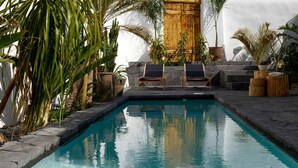 A pool area with two lounge chairs, surrounded by tropical plants and a wooden door. - Arithera Villa in Santorini (Santorini)