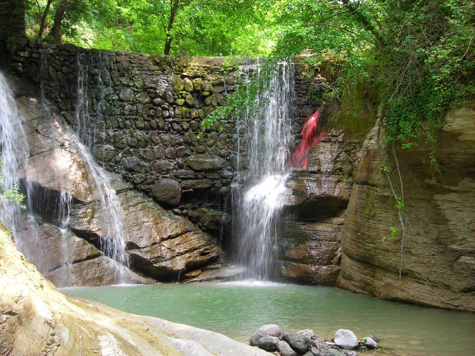 Ein natürlicher Wasserfall stürzt über einen Felsen in ein ruhiges Becken, umgeben von üppigem Grün.