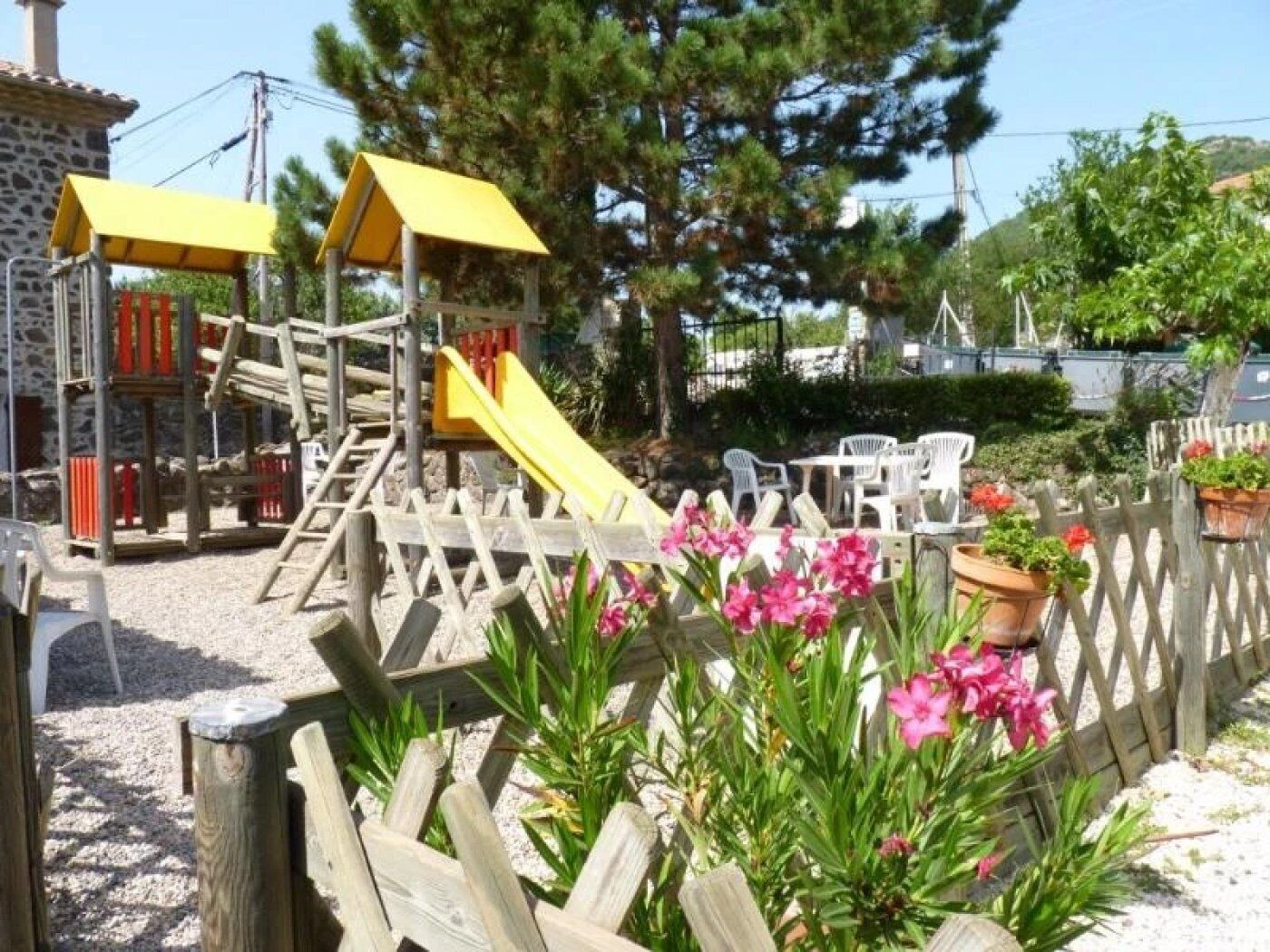 Une aire de jeux avec un toboggan et des structures d’escalade, entourée d’une clôture en bois et agrémentée de plantes en pot.
