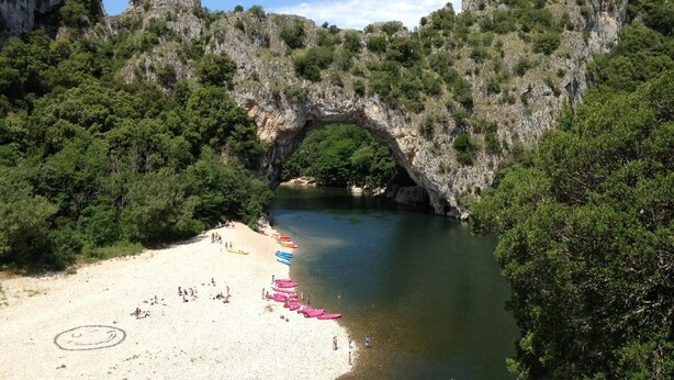 A beach with a natural rock arch formation, clear water, and a sandy shore with colorful kayaks.