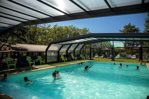 An indoor swimming pool with clear blue water, surrounded by green chairs and a covered area with a glass roof.