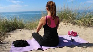 Una persona sentada sobre una toalla de playa mirando al océano, con un par de zapatos y un bolso cerca.