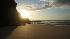 Una playa con una gran formación rocosa a la izquierda y el sol poniéndose sobre el océano.