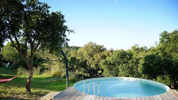 Una terraza de madera rodea una piscina azul cristalina con escaleras de metal, ubicada en un paisaje verde y exuberante.