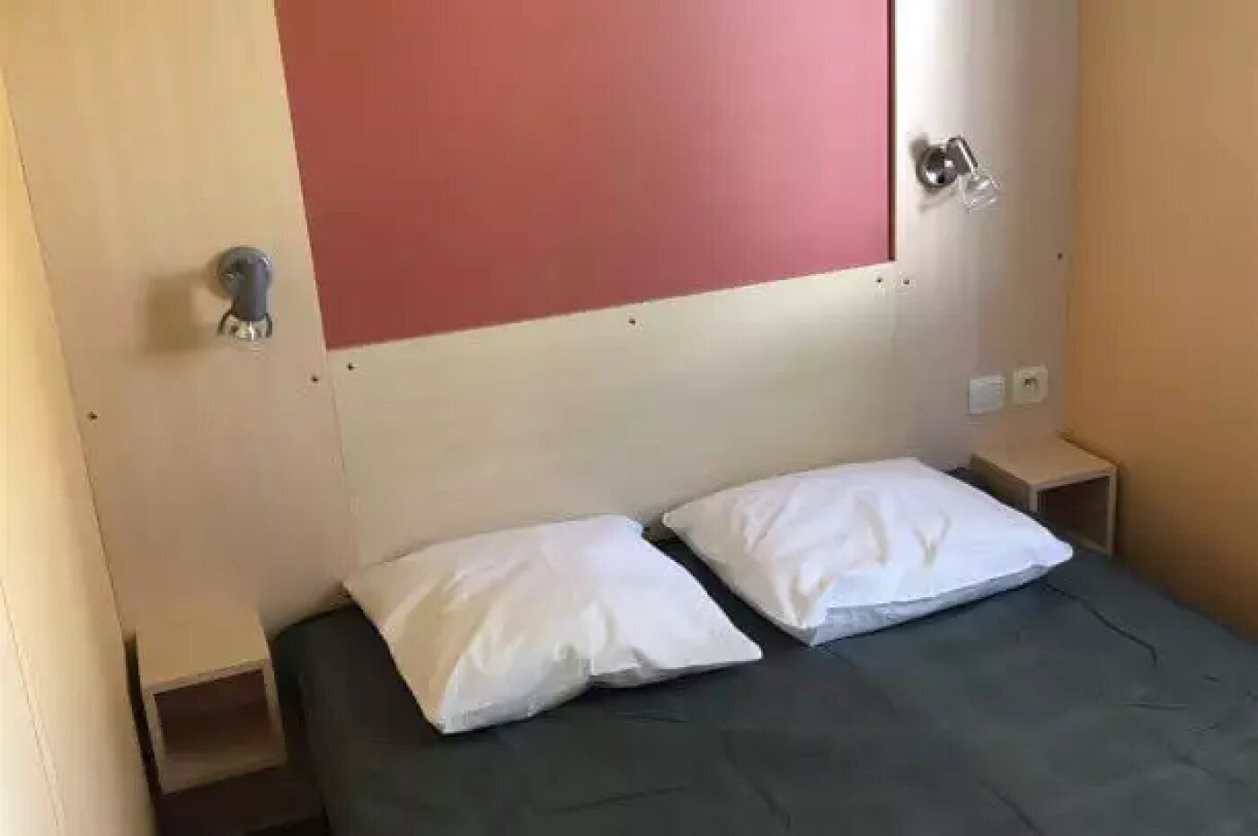 A small, simple room with a bed, two pillows, a nightstand, and a red panel on the wall.
