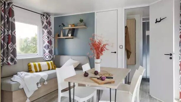 A modern living room with a sofa, a dining table with chairs, a bookshelf, and a window with patterned curtains.