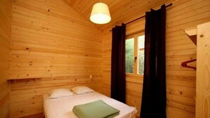 Ein Zimmer in einer Holzhütte mit einem Einzelbett, einer Pendelleuchte, einem Fenster mit Vorhängen und einer Tür mit einem darüber hängenden Handtuch.