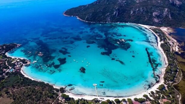 Ein Luftbild einer Küstenregion mit klarem türkisblauem Wasser, einem Sandstrand und einem gebirgigen Hintergrund.