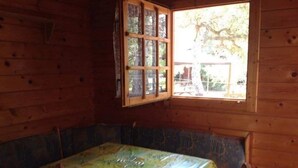 Ein Holzblockhaus-Interieur mit einem Esstisch, der mit einer blumigen Tischdecke bedeckt ist, einem Fenster mit Blick auf Bäume und einer Bank.