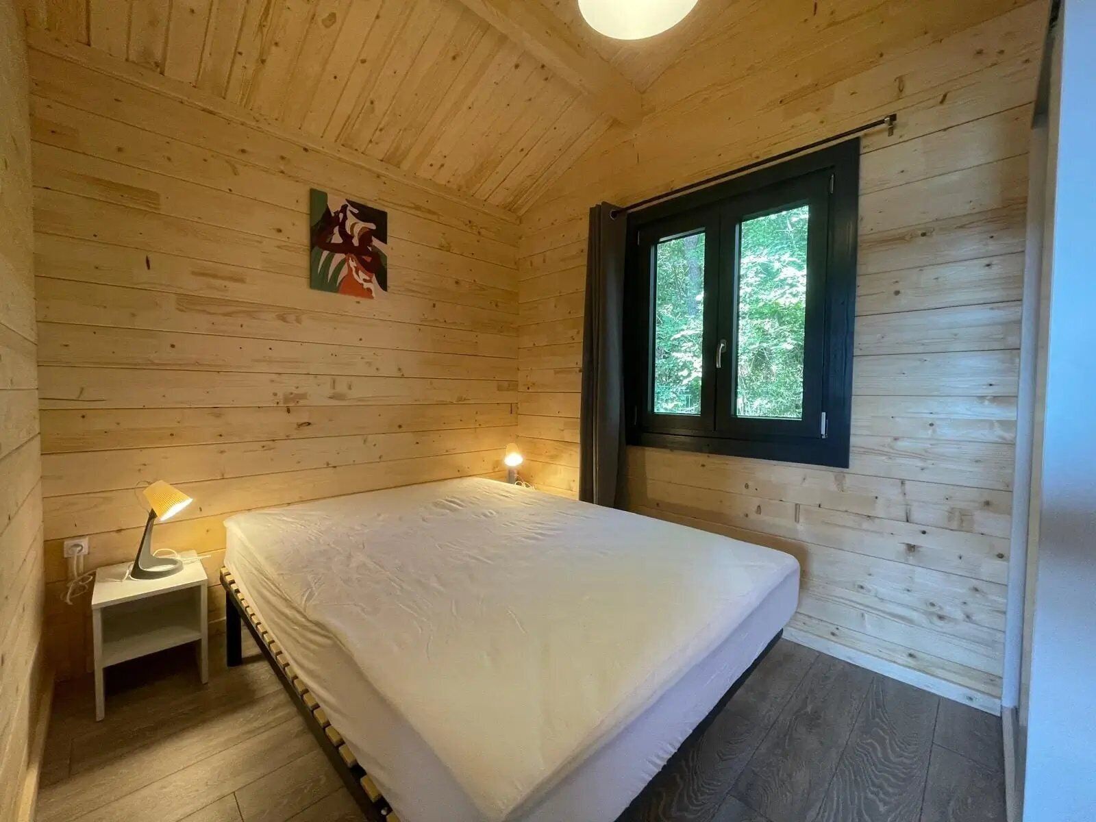 A wooden cabin room with a bed, a small bedside table, a window, and a painting on the wall.