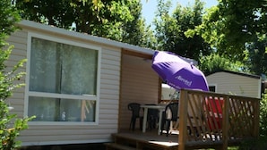 Ein Mobilheim mit überdachter Terrasse, einer Holzterrasse und einem violetten Airotel-Sonnenschirm.