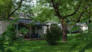 A single-story house with a covered porch, a small lawn, and a tree in the foreground. - ZEcamping 4 étoiles piscine couverte cottage 31m2 (Argentat)