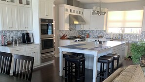 A modern kitchen with white cabinetry, a central island with bar stools, a dining area with a dark wooden table and chairs, and a large window with blinds. - Lake House (Lake Placid)