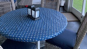 A covered patio with wicker furniture, a round table with a blue patterned tablecloth, and a wall clock. - Lake House (Lake Placid)