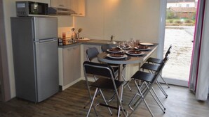 Cozinha compacta equipada com frigorífico, micro-ondas e fogão. Mesa de jantar posta com pratos e copos, e janela com vista para um edifício e vegetação.