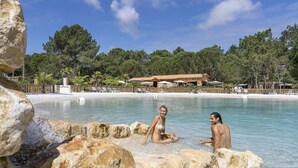 A pool area with large rocks in the foreground, a building with a red roof in the background, and a beach with white sand and trees. - ZEcamping 4* parc aquatique pet friendly club enfant locatif TV LV (Les Mathes)