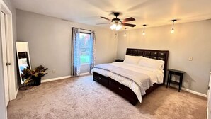 A bedroom with a bed, a ceiling fan, a small table, a nightstand, and a potted plant.