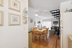 Ein Essbereich mit Holztisch und -stühlen, einem Deckenventilator und einer Küchenzeile mit Kühlschrank sowie einer schwarzen Treppe.