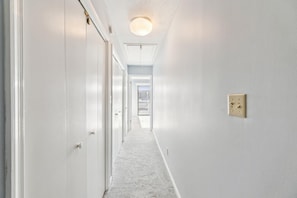 A hallway with white walls, a carpeted floor, a ceiling light, and a door at the end.