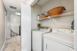 A laundry room with two washing machines, a dryer, and a shelf with baskets and bottles.