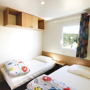 Ein Schlafzimmer mit zwei Einzelbetten, einem Holzregal darüber, einem Fenster mit Ausblick und einem Vorhang mit Blätermuster.