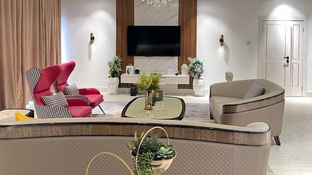 A modern living room with a large sectional sofa, a red armchair, a coffee table, and a TV mounted on the wall. - MacDonald Apartments A5  (Lagos)