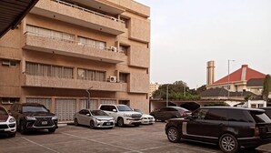 A parking lot with several cars, a multi-story building with balconies, and a red-roofed structure in the background. - MacDonald Apartments A5  (Lagos)