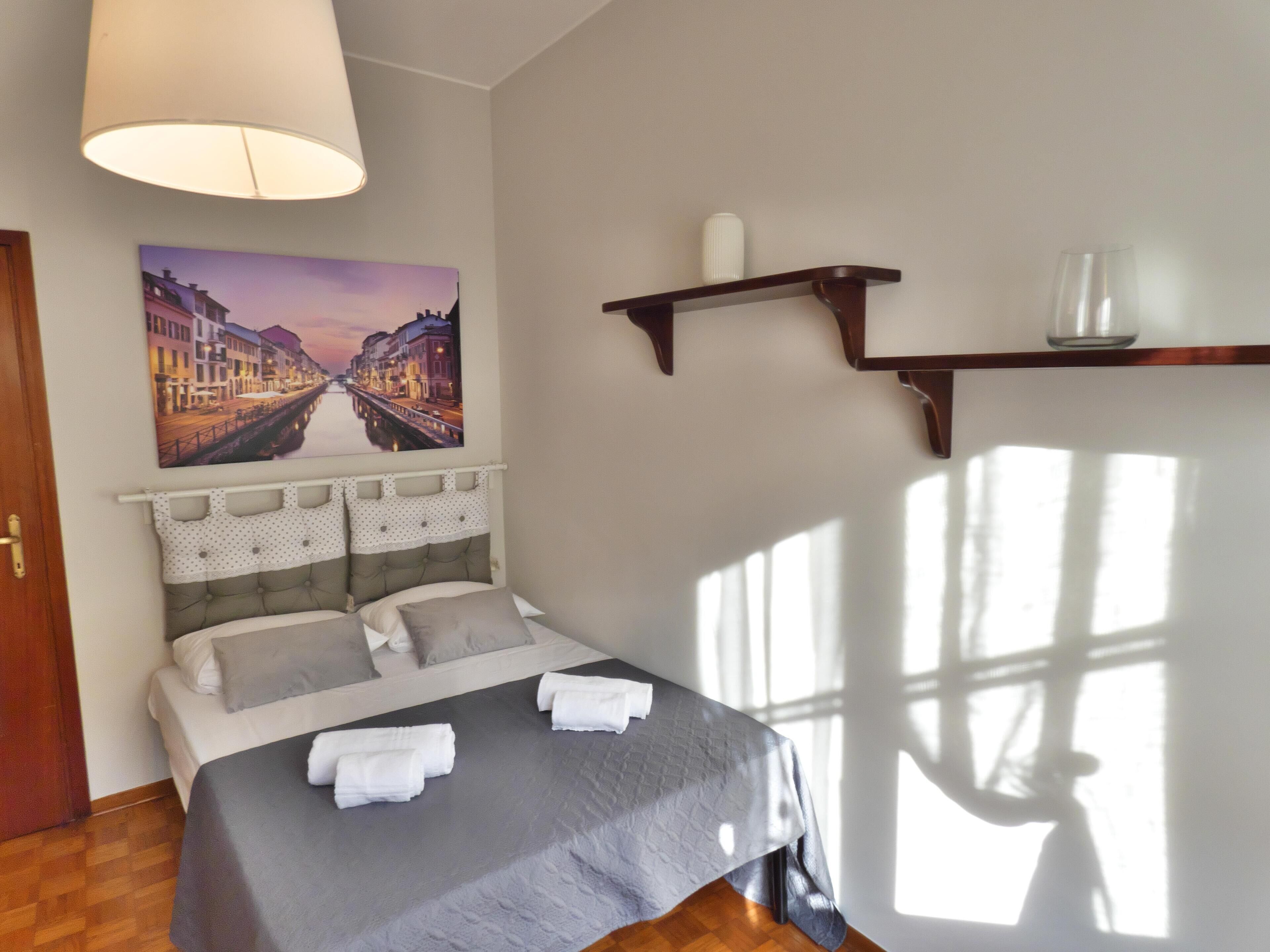 Een netjes opgemaakt bed met een grijze dekbedovertrek en witte kussens, een groot raam waar zonlicht schaduwen werpt, een wandplank met een vaas, en een stadslandschap schilderij boven het bed.