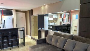 Una sala de estar moderna con un sofá gris, una zona de cocina con refrigerador y un box de ducha de vidrio.