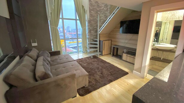 A modern living room with a large window, a sofa, a fireplace, and a kitchen area. - Saint Sebastian Flat 612 - Até 4 pessoas com hidro no centro (Jaragua do Sul)