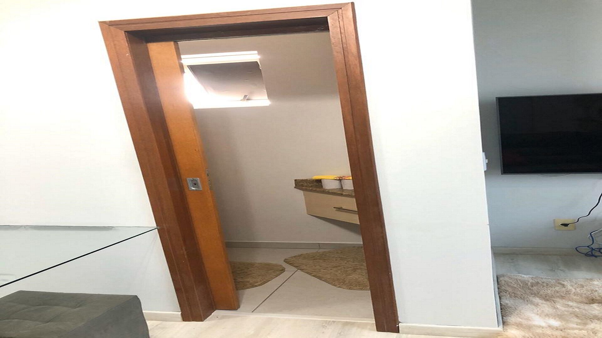 Quarto com porta de madeira, um pequeno espelho, luminária, televisão fixada na parede e piso coberto por carpete.