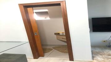 Quarto com porta de madeira, um pequeno espelho, luminária, televisão fixada na parede e piso coberto por carpete.