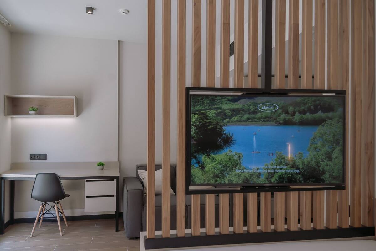 Een moderne hotelkamer met een grote televisie bevestigd aan een wand van houten panelen, een bureau met stoel, een bed en een klein plankje met een plant.