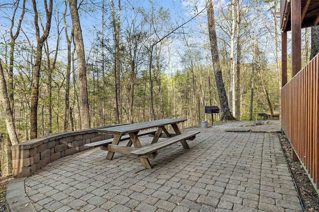 An outdoor picnic area with a wooden table and benches, surrounded by a brick wall and a wooden fence, set in a wooded area.