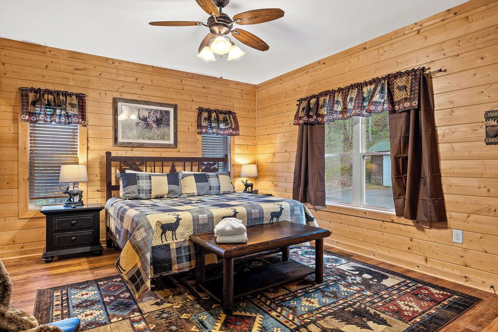 A cozy log cabin bedroom with a bed, two nightstands, a ceiling fan, and a large window with curtains.