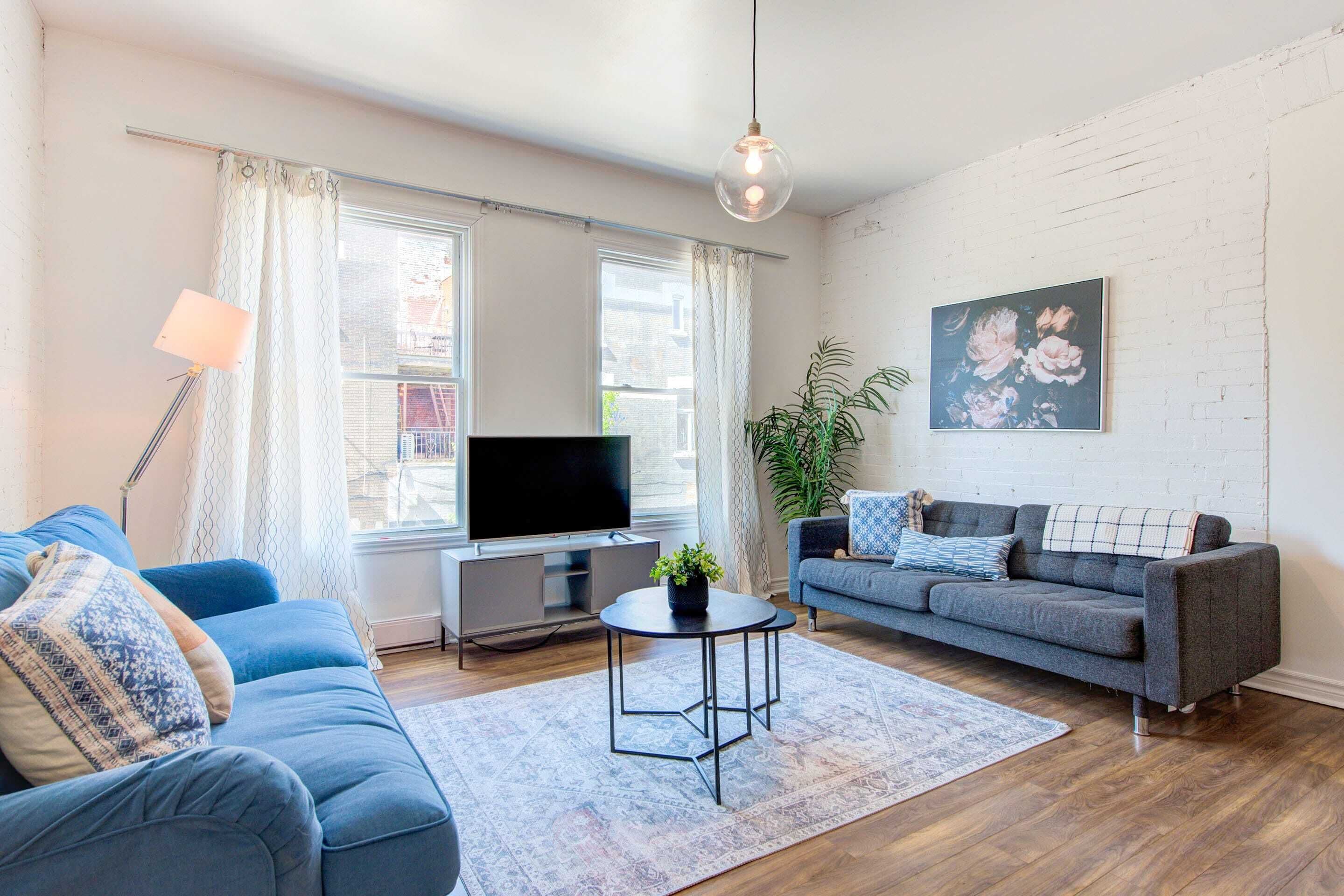 A modern living room with a blue sofa, grey sofa, a flat-screen TV, a small round table with a plant, and a large painting on the wall.