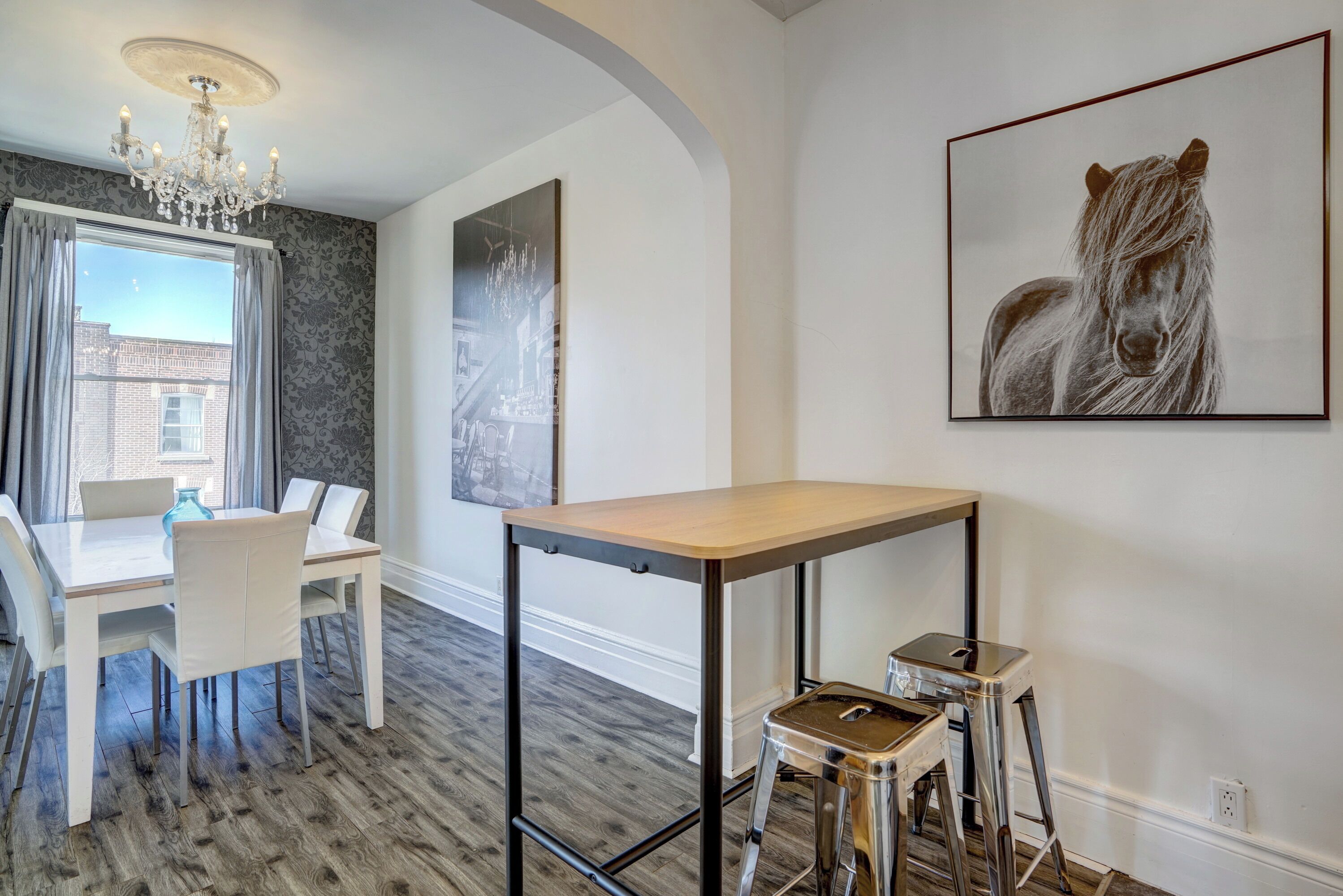 A dining area with a white table and chairs, a chandelier, and a cityscape painting on the wall.