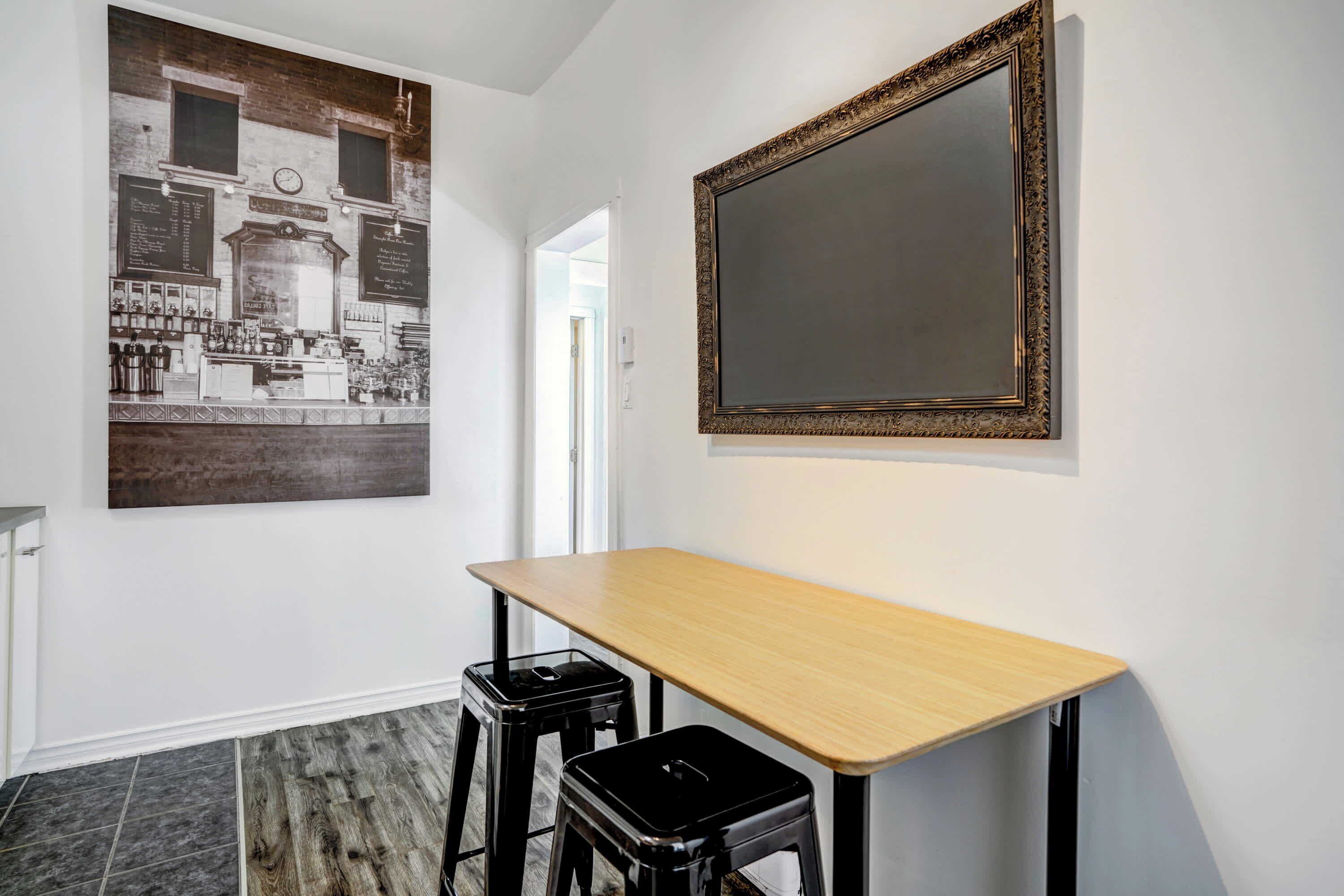 A room with a wooden table and two black stools, a large framed picture on the wall, and a tiled floor.