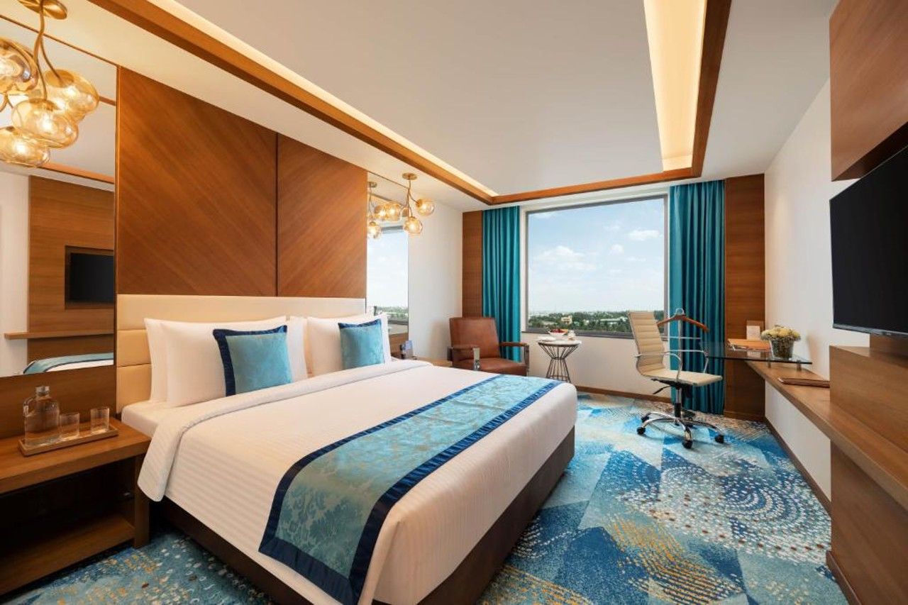 A hotel room with a large bed, a desk with a chair, a television, and a view of the city.