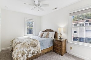 Ein Schlafzimmer mit Bett, Nachttisch, Lampe, Deckenventilator und einem Fenster mit Blick auf ein Haus.