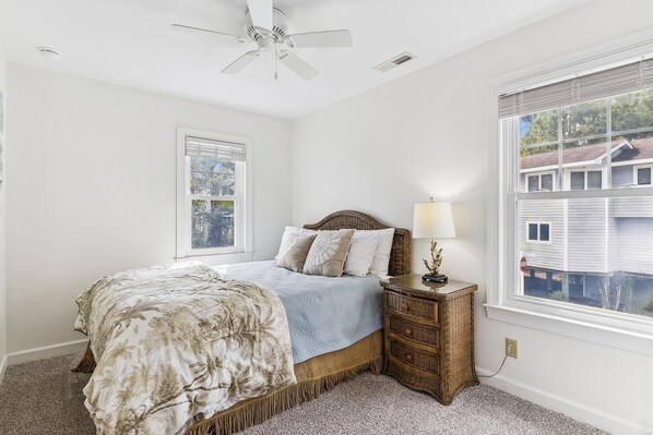 A bedroom with a bed, a nightstand, a lamp, a ceiling fan, and a window with a view of a house.