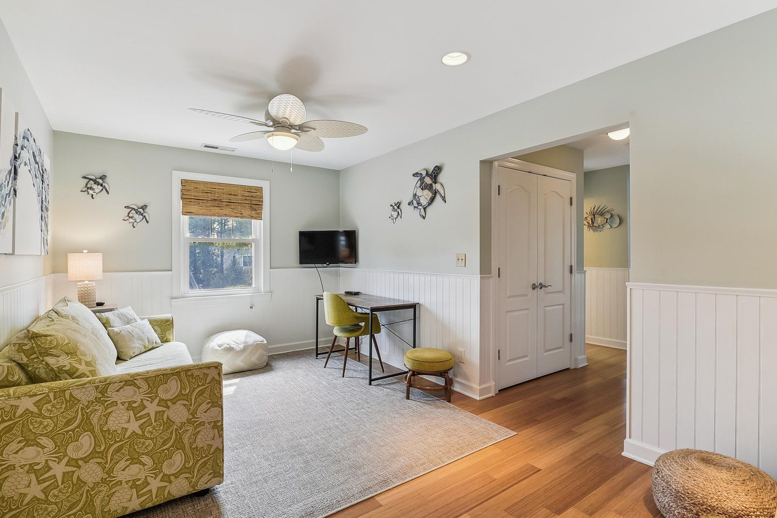 A room with a ceiling fan, a television, a desk with a chair, a sofa, and a door leading to another room.