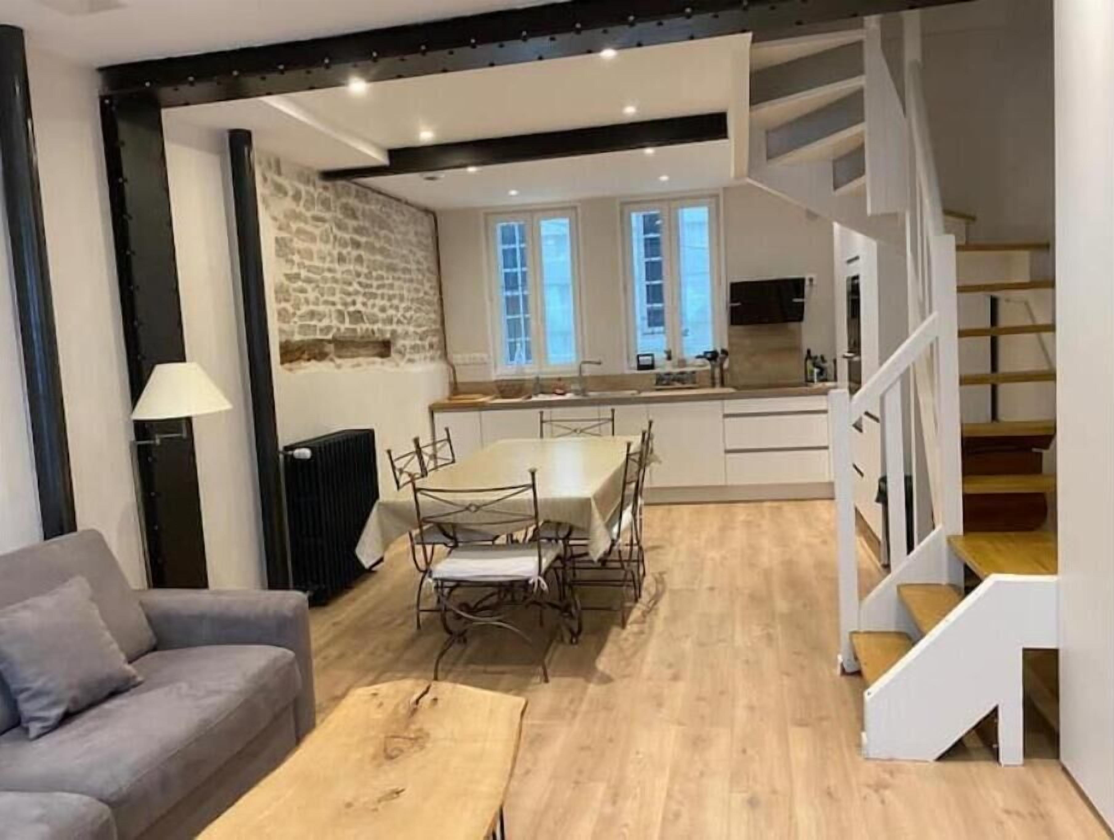 En moderne stue med en grå sofa, et træbord og et spiseområde med bord og stole. Et køkken er synligt i baggrunden med en stenvæg og en trappe, der fører op til en etage.