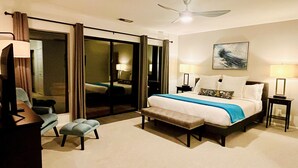 Una habitación de hotel con una cama grande, un escritorio, una silla, un banco, un ventilador de techo y un vidrio corredero que da a un balcón.